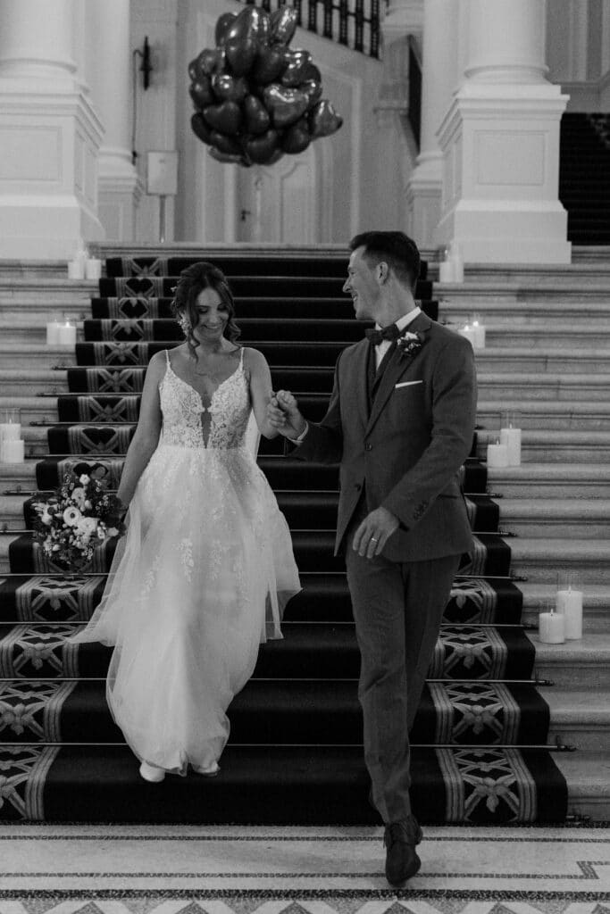 Braut und Bräutigam halten Händchen, während sie eine breite Marmortreppe im Palais Coburg hinabsteigen; die Braut trägt ein Spitzenkleid und hält in der linken Hand einen Blumenstrauß, im Hintergrund sind schwere Teppichläufer und große Säulen zu sehen.