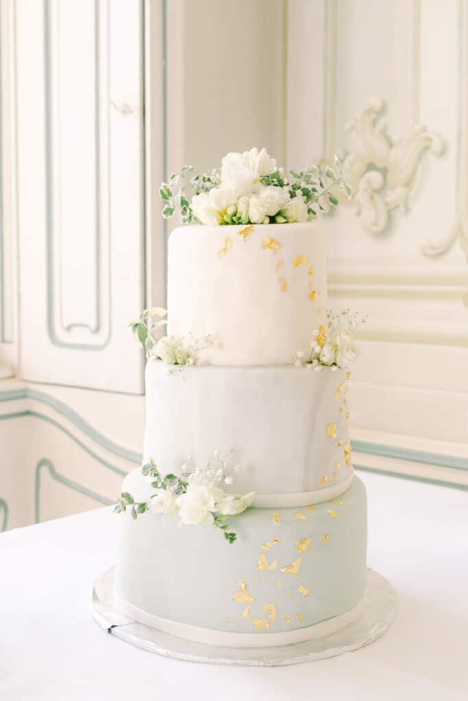 Elegante dreistöckige Hochzeitstorte mit Gold-Details und weißen Blüten – fotografiert von diehochzeitsfotografen.at bei einer Schlosshochzeit