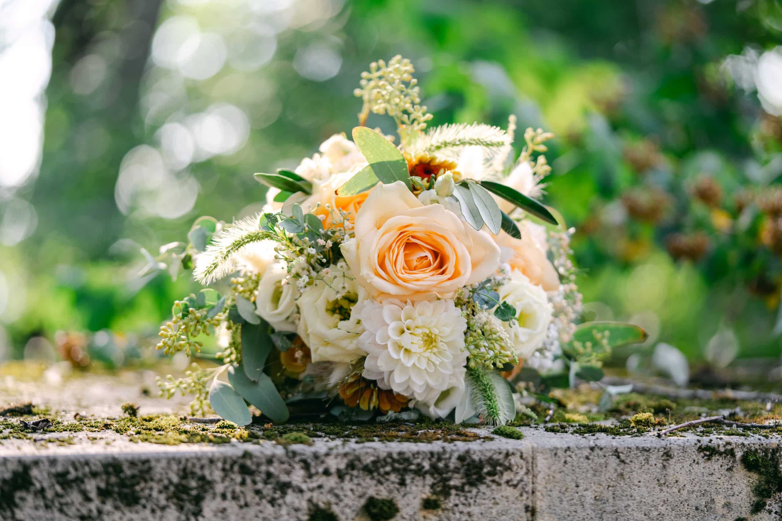 Brautstrauß mit apricotfarbenen Rosen, weißen Dahlien und Eukalyptus auf einer moosbedeckten Steinmauer im Schlossgarten von Schloss Vasoldsberg