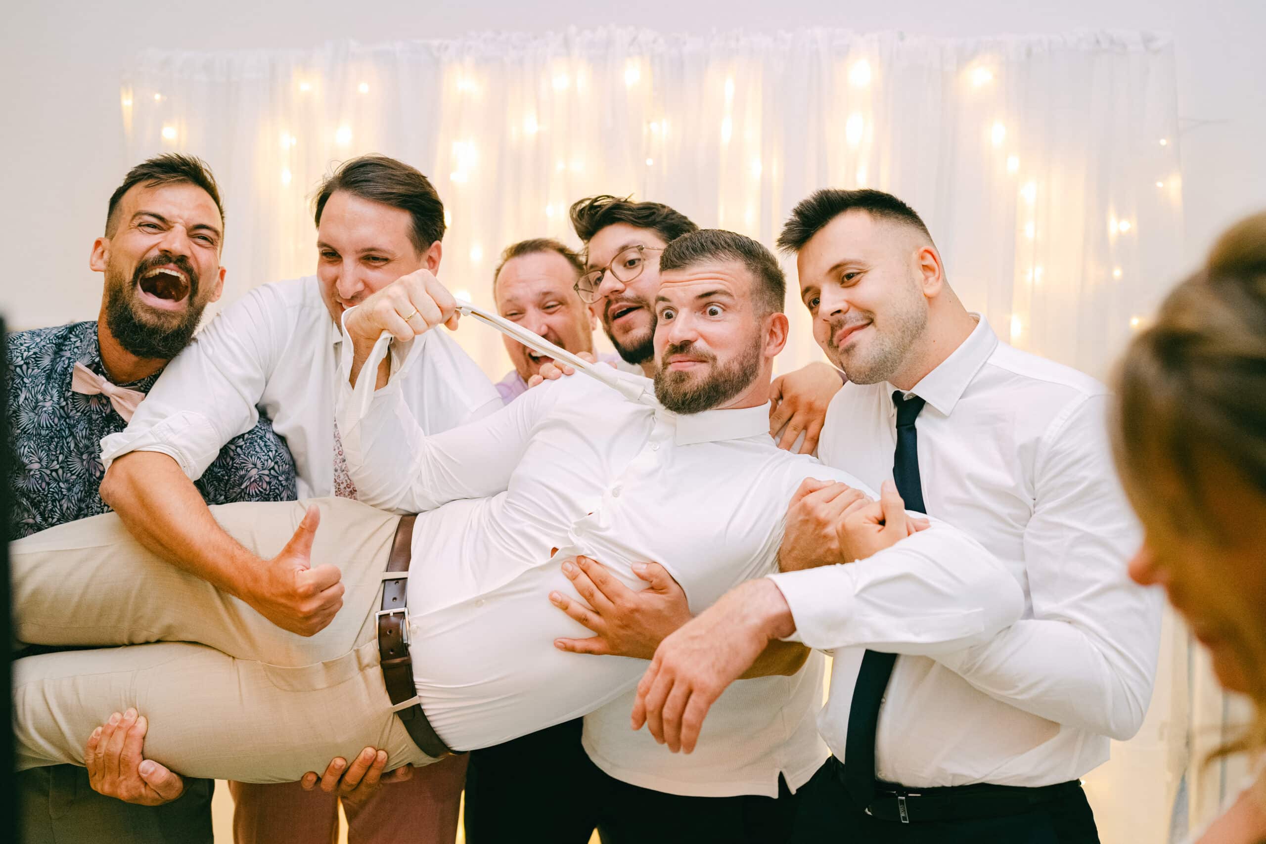 Gäste heben lachend den Bräutigam hoch vor einem Vorhang aus Lichterketten in der Fotobox