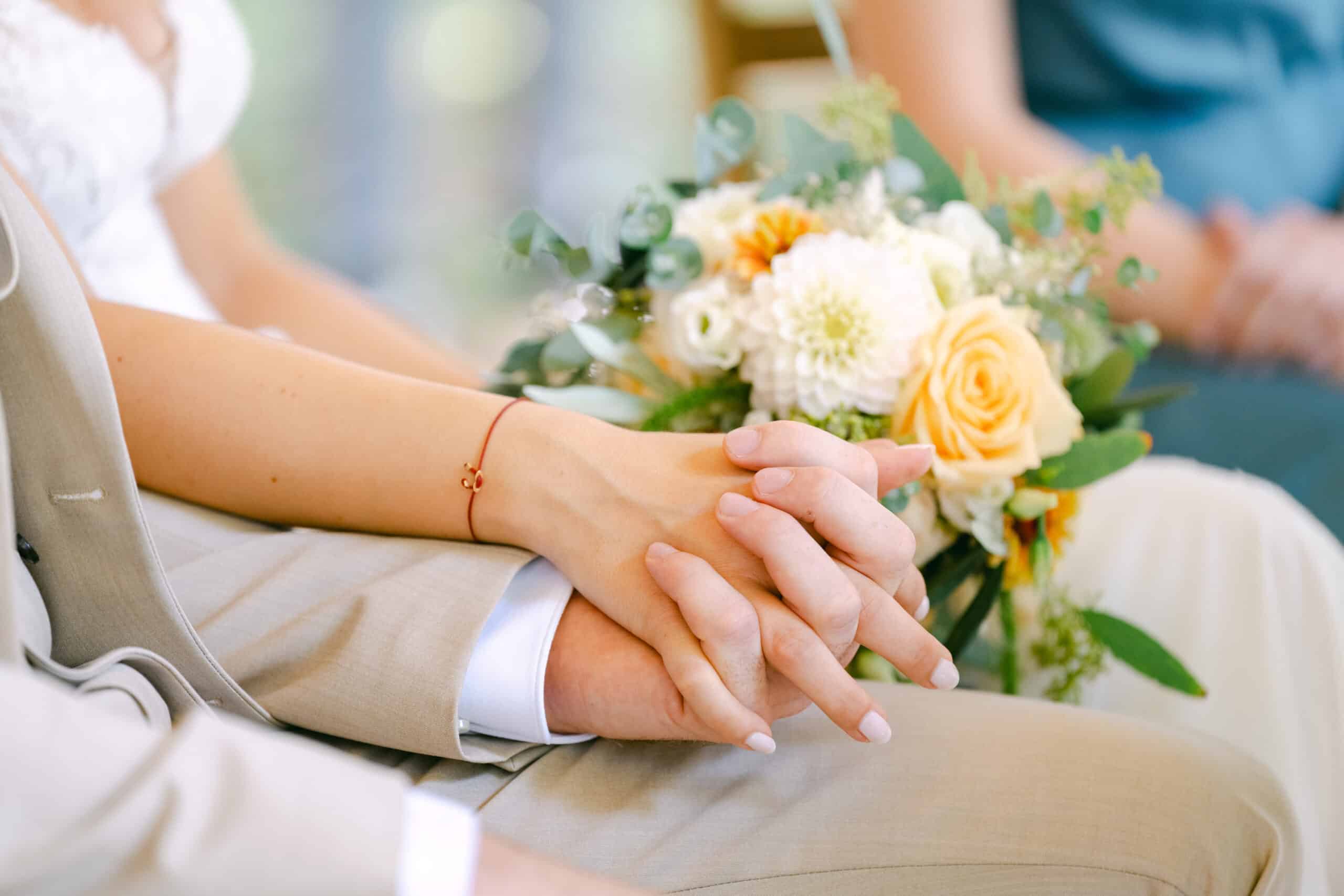 Braut und Bräutigam halten Händchen, im Hintergrund verschwimmt ein Brautstrauß aus weißen und gelben Blumen