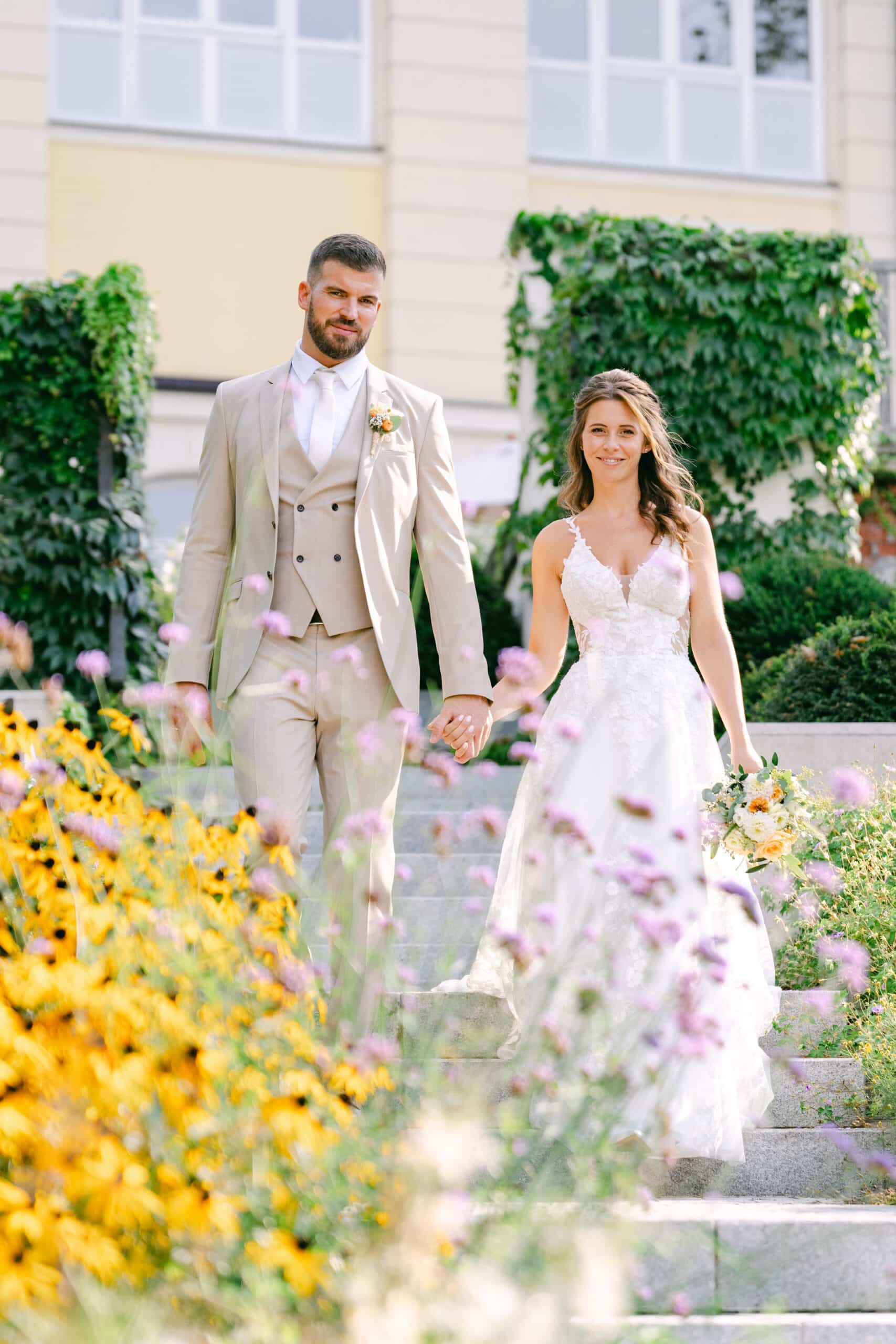 Braut und Bräutigam spazieren Hand in Hand durch den blühenden Schlossgarten von Schloss Vasoldsberg