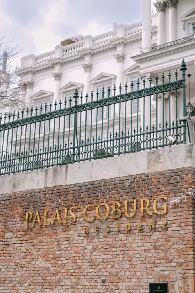 Goldene Lettern „PALAIS COBURG RESIDENZ“ auf einer roten Backsteinmauer, darüber ein grünes Ziergitter und im Hintergrund die weiße Säulenfassade des Palais Coburg.