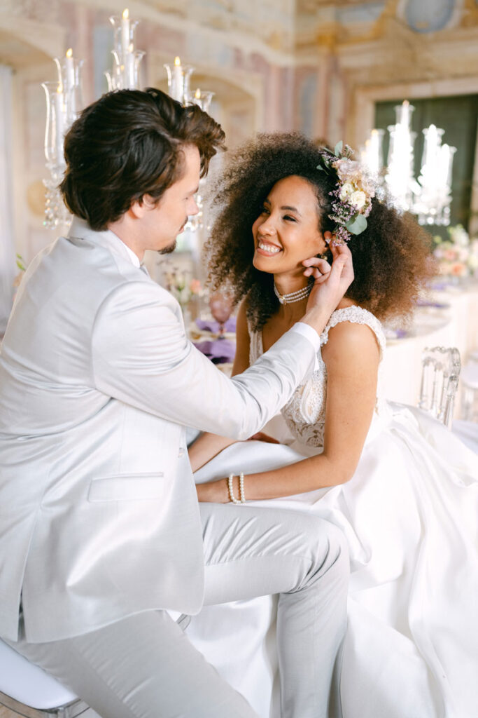 Ein kunstvoll gedeckter Hochzeitstisch mit lilafarbenen Stoffservietten, goldenen Besteckteilen und eleganten Kristallgläsern. Im Zentrum ein opulenter Kristallleuchter mit brennenden Kerzen, umrahmt von zarten Blumenarrangements in Rosa und Creme. Die Szene ist eingebettet in das stilvolle Ambiente des Schlosses Hagenberg – festgehalten von diehochzeitsfotografen für ein luxuriöses Editorial Wedding Shooting.