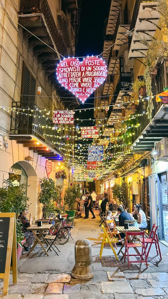 Belebte, festlich beleuchtete Gasse in Italien mit Herz-Leuchtschild und Straßencafés