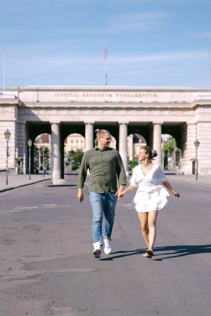 Paar hält Händchen und läuft vor dem Burgtor am Heldenplatz in Wien – strahlend blauer Himmel