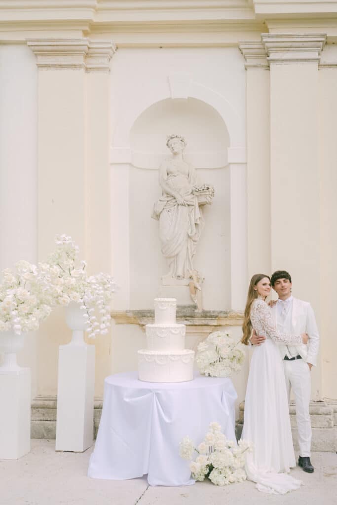 Braut und Bräutigam neben dreistöckiger Hochzeitstorte und weißen Blumenarrangements – Wedding Vienna diehochzeitsfotografen