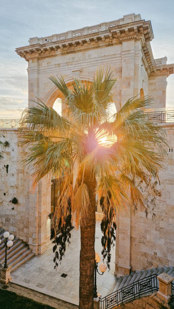 Sonnenstern strahlt durch historischen Steinbogen mit davorstehender Palme – Cagliari ist die Hauptstadt der italienischen Insel Sardinien Bastione Saint Remy