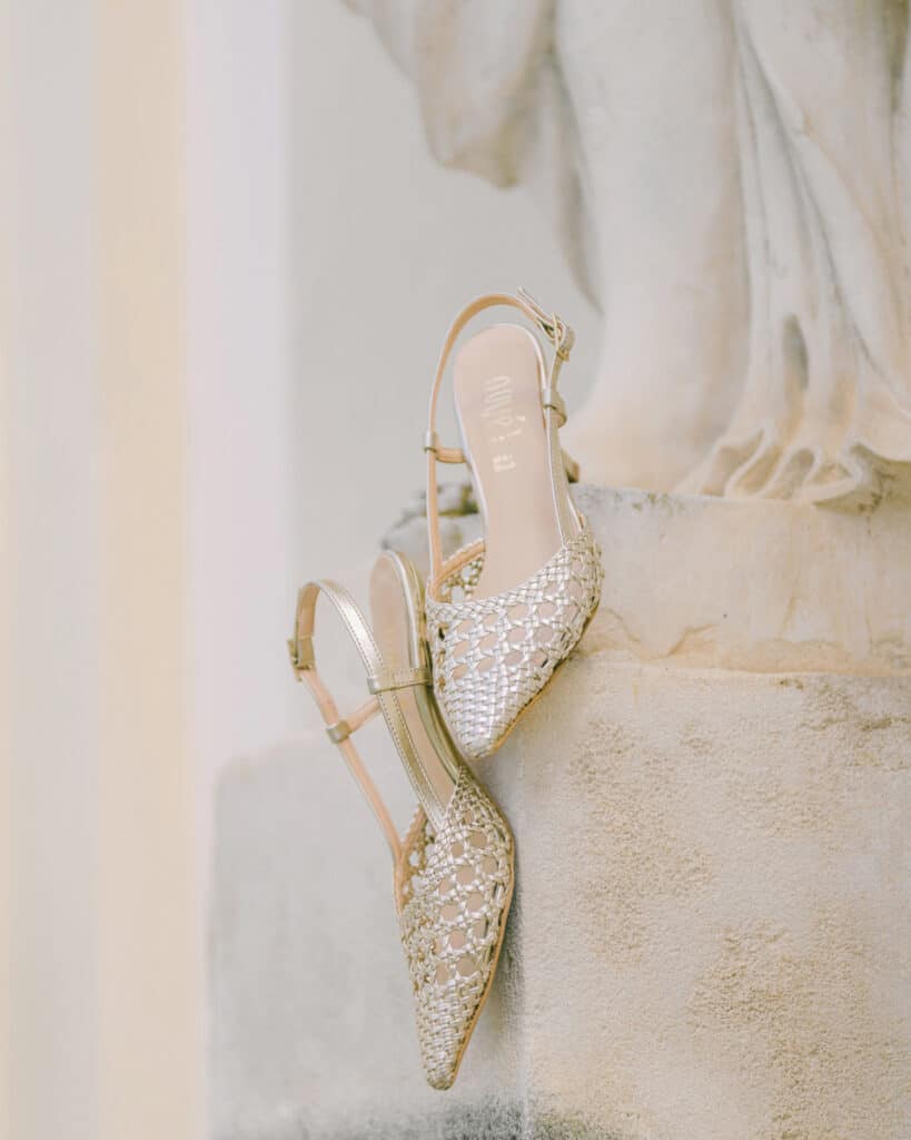Goldene geflochtene Brautschuhe lehnen dekorativ an einer Steinmauer – Detailaufnahme für Hochzeitsfotografie.