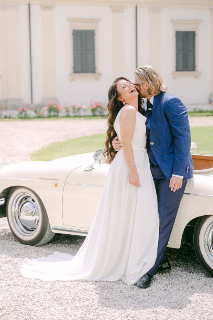 Hochzeitspaar vor Oldtimer in der Villa Mosconi Bertani – Braut lacht herzlich, während der Bräutigam sie küsst, festgehalten von die hochzeitsfotografen.